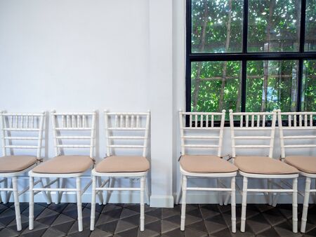 Row of clean white vintage wooden chairs with pads on modern stone tiles floor on white wall and glass window with green garden background.の写真素材