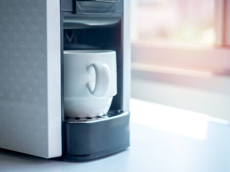 White ceramic coffee cup on coffee machine on white table near the window with sunlight in the morning with copy space.の写真素材