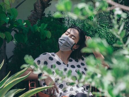 Beautiful Asian woman wearing protective face mask and polka dot shirt closing her eyes sitting on...
