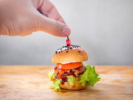 Homemade burger ready to eat. Hand put red cocktail pick on tasty mini homemade juicy burger with beef, cheese, fresh sliced tomato, green lettuce and caramelized onions on wood cutting board.の写真素材