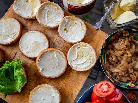 Process of making delicious homemade burgers. Top view of buns with butter, caramelized onions, fresh tomatoes on wood cutting board, prepared to make homemade burgers.の写真素材
