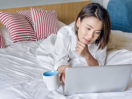 Beautiful Asian women with short hair wearing white shirt with white coffee cup and reading on laptop computer on wooden bed in white bedroom in the house.の写真素材