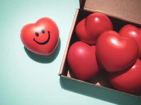 Red hearts with happy and smiling face on soft green background and many red ball foam with heart shape in brown paper craft box. Love, care, share, donation and Valentine Day Concept.の写真素材