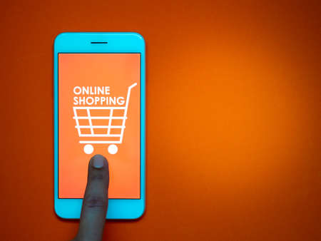 Online shopping concept. Words "Online Shopping" and shopping cart icon on orange background on smartphone screen with touching finger on orange background with copy space.の写真素材