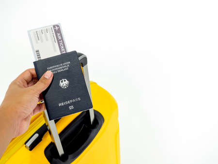 Hand holding passport and airline boarding pass ticket with yellow suitcase on white background with copy space. Blue passport of Germany and yellow luggage, travel concept.のeditorial素材
