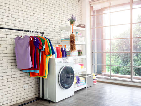Laundry room interior. Utility room with washing machine, cleaning equipment, home cleaners, clean wipes, hanging colorful shirts on clothesline on white brick wall background near large glass window.の写真素材