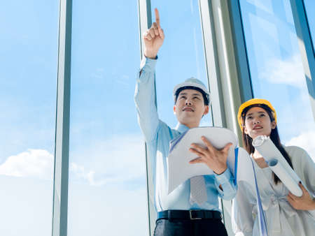 Asian architect Male and female inspecting construction work inside high-rise building near huge window glass on blue sky background. Couple engineer in hard hats holding blueprint in new building.の写真素材