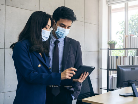 Asian business man and woman wearing suit and protective face masks using digital tablet, meeting and working together in office, new normal working lifestyle after pandemic lockdown.の写真素材