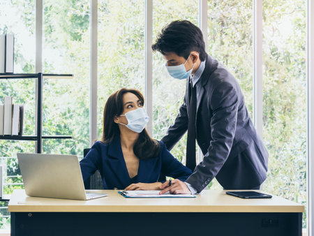 Asian business man and woman wearing suit and protective face masks using computer on desk, meeting and working together in office, new normal working lifestyle after coronavirus pandemic lockdown.の写真素材