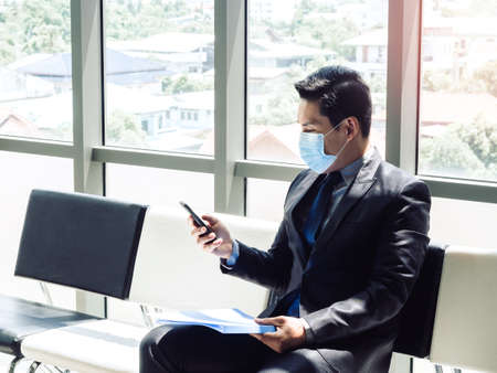 Asian businessman in suit wearing protective face mask using mobile phone while sitting in modern office building near huge glass window.の写真素材
