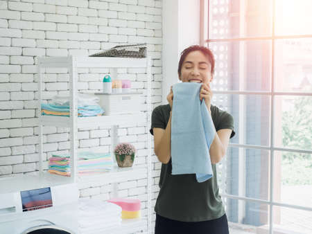 Beautiful young Asian woman housewife standing with smiling and smelling blue clean towel after washing from washing machine in laundry room with sunlight.の写真素材