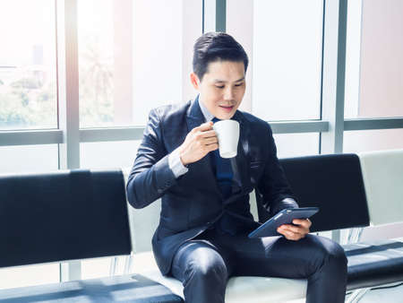 Good looking Asian businessman in suit watching tablet on his hand and drinking white cup of hot...