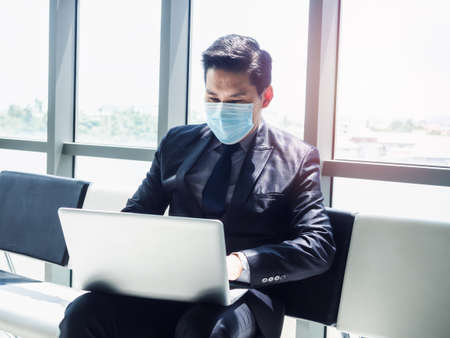 Asian businessman in suit wearing protective face mask using laptop computer on his lap while sitting in modern office building near huge glass window.の写真素材