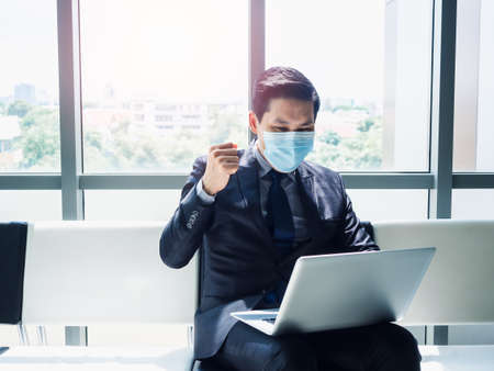 Asian businessman in suit wearing protective face mask raised his hand with happy and cheerful when he saw a laptop monitor on his lap while sitting in modern office building near huge glass window.の写真素材