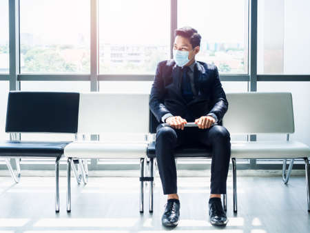 Asian businessman in suit wearing protective face mask sitting with his resume on his lap and waiting for job interview near huge glass window. Job Interview and unemployment concept.の写真素材
