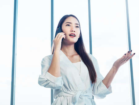 Young pretty Asian woman wearing formal white shirt talking on the phone call near huge glass window in office. Portrait of beautiful business woman using mobile phone in conversation.の写真素材