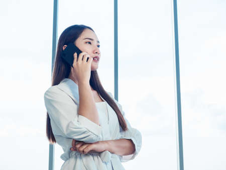 Young pretty Asian woman wearing formal white shirt talking on the phone call near huge glass window in office with copy space. Portrait of beautiful business woman using mobile phone in conversation.の写真素材