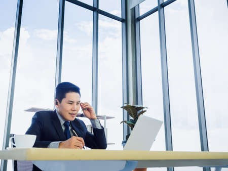 Asian businessman wearing suit with headset making notes and using laptop computer on desk in office near huge glass window and sky background. Conference call and customer support operator concept.の写真素材