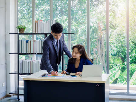 Asian business woman and businessman wearing suit working together in office. Young male employee standing pointing on chart for consult jobs with female boss while she working with computer on desk.の写真素材