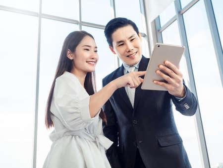 Business couple working together. Asian businessman in suit and smiling young woman pointing at digital tablet while both watching at screen on glass window background.の写真素材