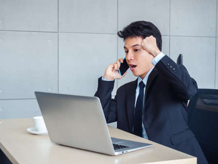 Young handsome Asian businessman in suit raising fist with exciting and shocking while calling with his mobile phone and looking laptop computer on desk in office with winner feeling and celebrating.の写真素材