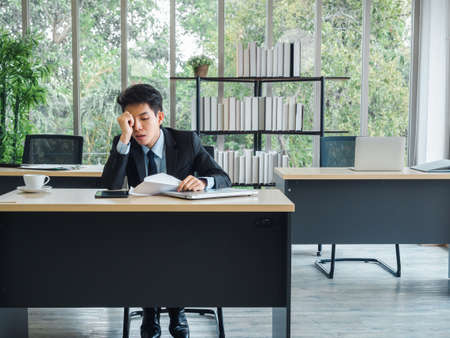Young Asian businessman receiving bad news with dismissal letter, tired, stressed and sad sitting with absent-minded on his desk in office. Frustrated worker mental health problems.の写真素材