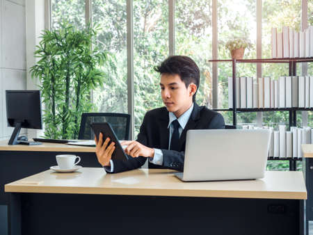 Young handsome Asian businessman in suit and tie sitting and reading news on digital tablet and working with laptop computer and white coffee cup on his desk in office in the morning with sunlight.の写真素材