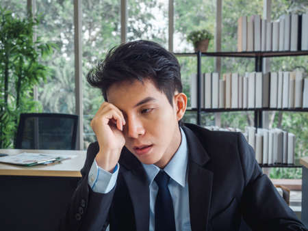 Young Asian businessman in suit with problems, tired, stressed and sad boring sitting with absent-minded on his desk in office. Frustrated worker mental health problems and burnout syndrome concept.の写真素材