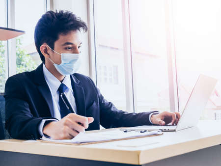 Young handsome Asian businessman in suit wearing face mask working on laptop computer and writing and checking business plan on chart while sitting in office near huge glass window with copy space.の写真素材