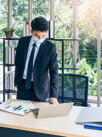 Young handsome Asian businessman in suit wearing protective face mask standing and working on laptop computer and checking business plan on chart in office near huge glass window, vertical style.の写真素材