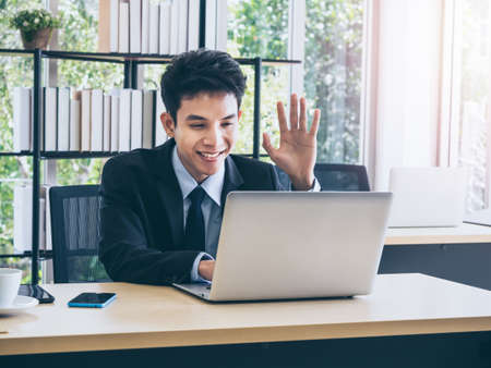 Young smiling Asian businessman in suit greeting someone, wave to camera on laptop computer have video conversation online while working on desk in office near huge glass window with sunlight.の写真素材
