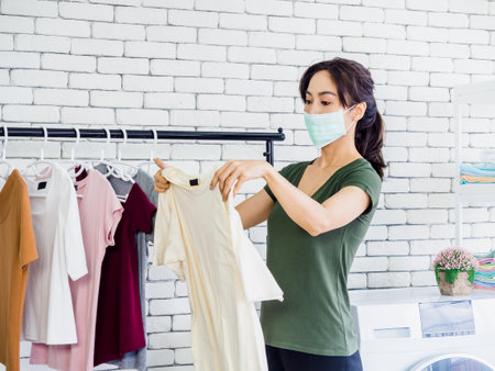 Young beautiful Asian woman, housewife wearing casual cloth and protective face mask holding shirt,...