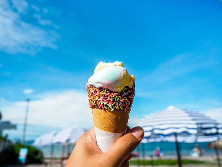 Ice cream in hand on beach and blue sky background. Rainbow color ice cream in cone with chocolate...