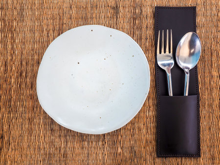 Ceramic plate. Empty white freeform shaped porcelain dish with stainless spoon and fork in leather case on mat, top view.の写真素材