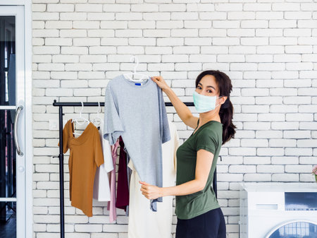 Young beautiful Asian woman, housewife in casual wearing protective face mask hanging dry shirt with hanger on clothesline after washing near wash machine in laundry room on white wall background.の写真素材