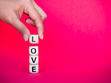 Love word written on wood block on pink red background with copy space, minimal style. Hand holding L to put on Love, text is written in black letters on wooden cubes.の写真素材