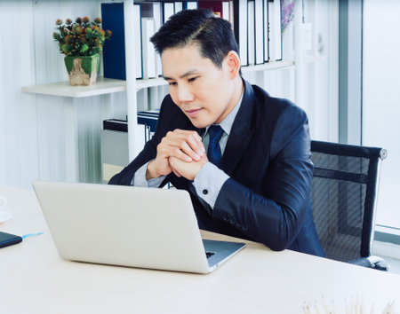 Handsome Asian businessman in suit concentrate watching on screen while working with laptop computer on desk in modern office container house near book shelf.の写真素材