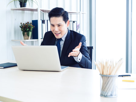 Handsome Asian businessman in suit video call, explaining to colleague through laptop computer in meeting online working, video conference. Social distancing or telecommunication work concept.の写真素材