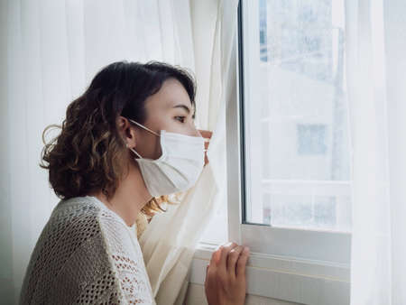 Beautiful lonely Asian woman wearing medical face mask looking out the window, stay at home for self-quarantine. Stay safe concept, prevention COVID-19 or Coronavirus pandemic situation.の写真素材
