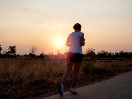 Young woman running on rural concrete road on beautiful bright sunset sky background near summer...