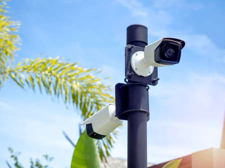 White CCTV cameras on the black pole in the garden near the building on blue sky background. Outdoor security CCTV camera surveillance.の写真素材