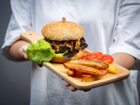Delicious fresh homemade burger set. Woman wearing casual white t-shirt holding and showing tasty homemade hamburger with french fries and sliced tomatoes on wooden tray or wood cutting board.の写真素材