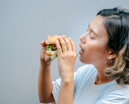 Delicious fresh homemade burger. Beautiful happy Asian woman short hair wearing casual white t-shirt holding and eating tasty homemade hamburger isolated on grey background.の写真素材