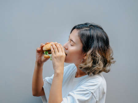 Delicious fresh homemade burger. Beautiful happy Asian woman short hair wearing casual white t-shirt holding and eating tasty homemade hamburger isolated on grey background.の写真素材