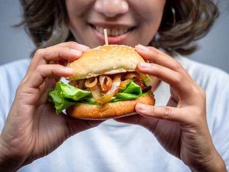 Delicious fresh homemade burger. Closeup beautiful happy Asian woman short hair wearing casual white t-shirt holding and eating tasty homemade hamburger isolated on grey background.の写真素材