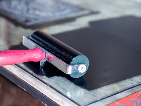 Close-up printmaking roller with red handle on black relief ink color on the glass in art studio with copy space. Linocut, woodcut, etching and silkscreen art printing step process.の写真素材