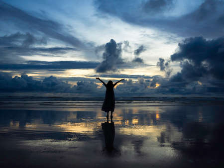 Beautiful sunset sky seascape background with silhouette of happy woman raising hands up on wet sand beach with reflection, dark sea and the vast sky background.の写真素材