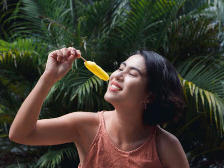 Woman eating ice cream. Happy beautiful Asian woman wearing casual pink tank top holding yellow ice cream on green tropical palm leaves background, outdoor.の写真素材