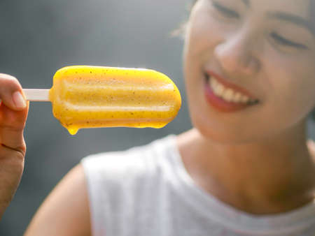Close up yellow ice cream in beautiful happy Asian womanâs hand wearing casual white sleeveless shirt, outdoorsの写真素材