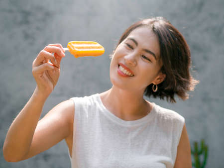 Yellow ice cream in beautiful happy Asian womanâ€™s hand wearing casual white sleeveless shirt,...